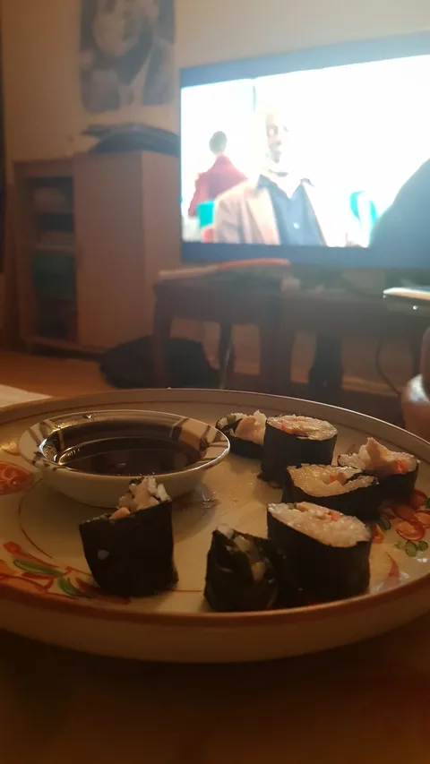 The image shows a cozy indoor setting. In the foreground, there is a plate with several pieces of sushi, including rolls wrapped in seaweed, filled with rice and what appears to be vegetables and fish. Beside the sushi is a small dish filled with soy sauce for dipping. The plate itself has a decorative floral design.
In the background, a television is on, showing a scene with two people in conversation. The image on the TV is slightly blurred, making it difficult to discern details about the show or movie. The lighting in the room is warm and soft, creating a relaxed atmosphere, typical of a casual meal at home. The furniture, such as a wooden table and a cabinet, adds to the homey ambiance. The television serves as a focal point, suggesting this is a moment of leisure, possibly during a personal viewing session.