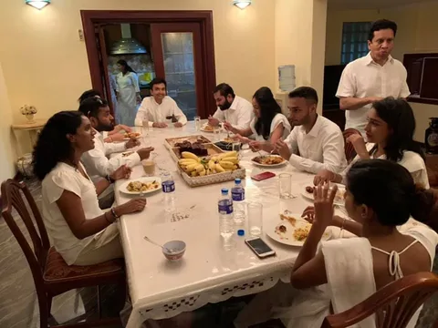 1) Rich description: The image captures a dining scene in an indoor setting, with a group of people seated around a large rectangular table. The table is covered with a white tablecloth and is laden with plates of food, bottles of water, and a central platter of bananas and cake. The individuals are dressed in white, suggesting a formal or coordinated gathering. At the far center end of the table are US Army Captain Hashani Hettiarachchi and Lehan Edirisinghe, both engaged in conversation and eating. Standing to the right, also dressed in white, is Saroj Perera, who is holding a plate and appears to be eating while standing. The mood is casual and sociable, with people interacting and enjoying their meals. The lighting is warm, casting a cozy ambiance over the dining area. In the background, a kitchen area is partially visible through a doorway, with another person standing inside. Overall, the scene conveys a sense of camaraderie and conviviality among the participants.
2) On-screen text block:
- Title: "..."
- Channel / profile: "..."
- Site / app: "..."
- Captions / subtitles: "..."