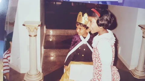 ```
In this image, two children are dressed in costumes, suggesting a festive or playful occasion, possibly a party or school event. The child identified as Lehan Edirisinghe is wearing a regal costume, complete with a golden crown, a purple cloak, and a black outfit adorned with gold buttons. He holds a piece of paper or card, possibly part of his costume or a prop. Standing beside him is Wagisha Sumathipala, wearing a floral dress with a white lace collar and a red headband, suggesting a more formal or thematic attire. Both children are positioned in front of a staircase with marble steps, flanked by decorative pillars, contributing to the setting's elegant ambiance. The lighting is warm, enhancing the festive and joyful mood. Their expressions are joyful, indicating they are enjoying the event or moment.