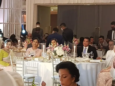 The image depicts an indoor formal event, likely a banquet or a wedding reception. The setting is elegant, with guests seated at round tables adorned with white tablecloths, floral centerpieces, and tall candle holders. Among the attendees are Shanika Edirisinghe, Shavendra Silva, and Thilanga Sumathipala, as indicated by the hint. The guests appear to be in formal attire, with women in traditional and elegant dresses and men in suits and ties.
The atmosphere seems convivial, with people smiling and engaged in conversation. The lighting is warm, highlighting the decor and creating a festive ambiance. Some attendees are captured mid-conversation, while others are looking towards the camera. In the background, staff members are visible, some wearing masks, suggesting a level of formality and protocol. The overall mood is sophisticated and celebratory, typical of a high-profile social gathering. The image conveys a sense of community and festivity.