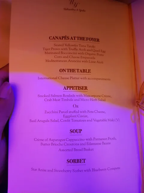 **Rich Description:**
This image shows an elegant menu from what appears to be a formal event, possibly a wedding or similar gathering, as indicated by the names "Udhantha & Yuki" at the top. The menu is presented on a cream-colored card with a subtle decorative border, and the text is in a sophisticated font, enhancing the upscale feel. The lighting in the image has a warm tone, creating an intimate and inviting atmosphere.
The menu is divided into several sections: "Canapés at the Foyer," "On the Table," "Appetiser," "Soup," and "Sorbet." Each section offers a selection of gourmet dishes. Under "Canapés at the Foyer," items like Seared Yellowfin Tuna Tataki and Tiger Prawn with Truffle Aioli are listed, suggesting a focus on high-quality ingredients and intricate preparations. An "International Cheese Platter" is set to be on the table, adding to the luxurious experience.
The "Appetiser" section offers two options: Smoked Salmon Roulade with Mascarpone Crème, and a vegetarian alternative featuring Zucchini Parcel stuffed with Feta Cheese. The "Soup" section features a Crème of Asparagus Cappuccino with Parmesan Froth. For a palate cleanser, the "Sorbet" section lists a Star Anise and Strawberry Sorbet with Blueberry Compote. The attention to detail in the menu items reflects a keen emphasis on culinary artistry and presentation.
**On-screen Text Block:**
- Title: "Udhantha & Yuki"
- Key OCR Lines:
- "CANAPÉS AT THE FOYER"
- "Seared Yellowfin Tuna Tataki"
- "Tiger Prawn with Truffle Aioli and Quail Egg"
- "Marinated Bocconcini with Organic Pesto"
- "Corn and Cheese Empanada"
- "Mediterranean Arancini with Lime Aioli"
- "ON THE TABLE"
- "International Cheese Platter with accompaniments"
- "APPETISER"
- "Smoked Salmon Roulade with Mascarpone Crème"
- "Crab Meat Timbale and Micro Herb Salad"
- "OR"
- "Zucchini Parcel stuffed with Feta Cheese"
- "Eggplant Caviar"
- "Basil Arugula Salad, Confit Tomatoes and Vegetable Maki (V)"
- "SOUP"
- "Crème of Asparagus Cappuccino with Parmesan Froth"
- "Butter Brioche Croutons and Edamame Beans"
- "Assorted Bread Basket"
- "SORBET"
- "Star Anise and Strawberry Sorbet with Blueberry Compote"