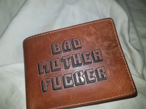 The image shows a close-up of a brown leather wallet lying on a white fabric surface, likely a bed or a couch. The wallet features prominently embroidered text in bold black letters that reads "BAD MOTHER FUCKER." The text is centered and stands out due to its contrasting color and the texture of the leather. The leather appears slightly worn, suggesting it might be well-used or intentionally distressed for aesthetic purposes. The stitching around the edges of the wallet is visible, and the overall lighting is even, providing a clear view of the texture and color of the materials. This style of wallet is reminiscent of a popular cultural reference, which adds a playful or bold statement to the item. The setting appears casual and relaxed, given the soft fabric background.