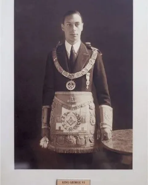 In this historical portrait, King George VI is depicted wearing formal Freemason regalia. The setting is a simple, indoor backdrop that highlights the solemnity and dignity of the occasion. King George VI stands upright, facing the camera, conveying a sense of authority and composure. He is dressed in a dark formal suit with a white dress shirt and a tie, over which he wears an elaborate Masonic apron featuring intricate embroidery and symbols, such as the square and compasses, and various other Masonic emblems. Around his neck, he wears a Masonic collar adorned with additional symbols and medals. The lighting is soft, casting subtle shadows that add depth to the image, while the sepia tone gives it a vintage, timeless quality. Below the portrait, there is a plaque with the text "KING GEORGE VI," identifying him explicitly. The overall ambiance of the photograph is formal and respectful, reflecting the historical significance of the subject.