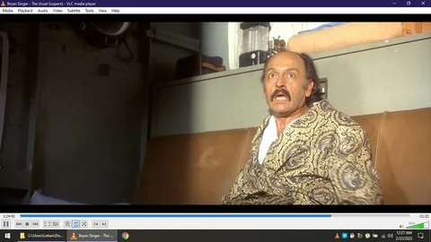 This image is a screenshot from a movie being played on VLC media player. The title at the top indicates it is from "The Usual Suspects," directed by Bryan Singer. The scene captures a man who appears to be sitting indoors, possibly in a train or a confined space due to the visible compartment and shelving. The man is wearing a patterned robe and has a surprised or startled expression on his face, with wide eyes and an open mouth. The lighting is somewhat dim, casting shadows on the wall behind him. The setting has a vintage feel, complemented by the muted colors and decor. The screenshot captures a moment of high tension or revelation, typical of the film's suspenseful style. The media player interface shows the timeline and controls at the bottom, indicating the video is near the end.