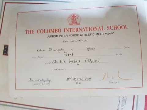 This image shows a certificate awarded by The Colombo International School for a Junior Inter House Athletic Meet in 2005. The certificate is primarily in white with a red border and text. At the top, the institution's name is prominently displayed in bold red letters: "THE COLOMBO INTERNATIONAL SCHOOL." Below, the certificate details indicate it is for a first-place finish in the Shuttle Relay (Open) event. Specific fields include placeholders for the recipient's name and house, which appear to be filled in with handwritten text. The document is signed by the Director of Sports and the Principal, with the date noted as 22nd March 2005. The certificate is likely printed on a standard sheet of paper, possibly framed or displayed against another document in the background. The overall composition suggests an official and formal recognition of athletic achievement within the school setting.