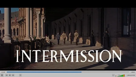 In this video frame from the film "Lawrence of Arabia," the scene is set in an expansive outdoor corridor of a historic building, likely representing a location in the film's Middle Eastern setting. The architecture features grand columns and arches, exhibiting a classical design with intricate details. Several individuals, dressed in period military uniforms and civilian attire, are walking through the corridor, casting long shadows on the tiled floor due to the bright sunlight. The atmosphere conveys a sense of historical grandeur and military formality. A British flag can be faintly seen in the background, hinting at the colonial context of the setting. The word "INTERMISSION" is prominently displayed across the bottom of the image, indicating a break in the film's narrative. The lighting is warm, contributing to the majestic and timeless feel of the scene, fitting the epic nature of the film.