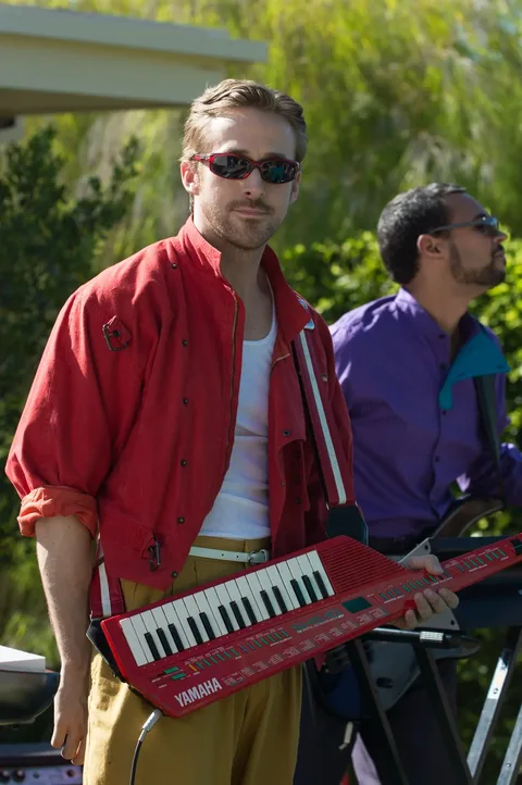 ```
Ryan Gosling is depicted in a scene from "La La Land," holding a red Yamaha keytar, adding a vibrant pop of color to the image. He is dressed in a casual yet striking ensemble featuring a red jacket over a white tank top and yellow pants, with rolled-up sleeves giving a relaxed vibe. His demeanor is cool and confident, accentuated by dark sunglasses, adding to the charismatic aura. The background features greenery and part of a building, suggesting an outdoor setting during the day. Another musician is partially visible, wearing a purple shirt and playing a guitar, contributing to the performance atmosphere. The lighting is bright and natural, highlighting the lively tone of the scene.