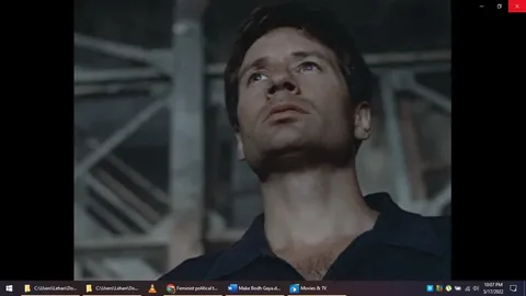```
**SECTION 2:**
The image is a still from a film or television sequence viewed in the "Movies & TV" application. The focus is on a man looking upwards thoughtfully, set against a backdrop of industrial-looking scaffolding or metal structures. The lighting is dim, suggesting an indoor or shaded environment, with shadows accentuating his serious and contemplative expression. He wears a dark shirt, and his posture is upright, conveying a sense of determination or reflection. The composition centers on his face, emphasizing his upward gaze and the mood of introspection or anticipation. The scene has a cinematic quality, with muted colors matching the somber tone. The bottom of the image shows parts of the desktop or application interface, including file paths and other open files or applications.