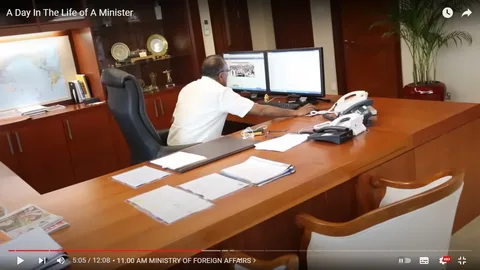 A man, referred to as K Shanmugam, is seated at a large wooden desk in an office setting. He is dressed in a white shirt and is focused on working at a dual-monitor computer setup. The room is neatly organized, with several papers and documents spread across the desk. On the desk, there are multiple telephones, suggesting a busy and high-level workplace environment typical of a ministerial office.
In the background, there is a wooden cabinet displaying various awards and trophies, indicating a place of importance and prestige. A world map is visible on the wall, adding to the official ambiance of the space. The lighting is bright and even, contributing to a professional atmosphere. This setting is part of a video titled "A Day In The Life of A Minister," and the specific time mentioned in the subtitle is "11:00 AM MINISTRY OF FOREIGN AFFAIRS," situating the scene within governmental or diplomatic activities.