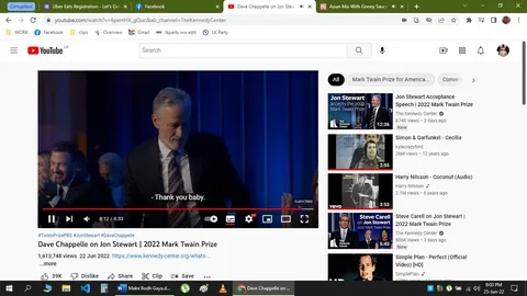 The image is a screenshot from a YouTube video titled "Dave Chappelle on Jon Stewart | 2022 Mark Twain Prize," hosted on The Kennedy Center's channel. The scene captures an indoor event, likely an award ceremony, since it involves the Mark Twain Prize. The video thumbnail shows a person on stage, presumably Jon Stewart, as suggested by the hint. The individual is dressed in a formal suit, standing in front of a dark blue curtain backdrop, suggesting a formal and elegant setting. The atmosphere appears celebratory, with an audience visible in the background, including people applauding. The text "Thank you baby" is part of the captions, indicating a moment of gratitude or affection. The video seems to focus on Jon Stewart, who is a central figure in the event. The YouTube interface is visible, with various video recommendations related to Jon Stewart and other similar topics on the right sidebar.