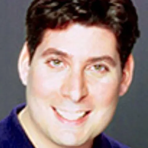 The image is a portrait-style headshot of a person identified as Jonathan Uman. The photo is a close-up, capturing the subject from the shoulders up against a plain, neutral background. Jonathan Uman has short, dark hair and is smiling directly at the camera, conveying a warm and approachable demeanor. The lighting is soft, highlighting his facial features without harsh shadows, which suggests a professional studio setting. He is wearing a dark-colored shirt, which contrasts with the lighter, possibly gray, background, allowing his face to be the focal point of the image. The overall tone of the photograph is bright and clear, emphasizing a friendly and professional appearance. There are no visible logos or text in the image, focusing entirely on Jonathan Uman's expression and presence.