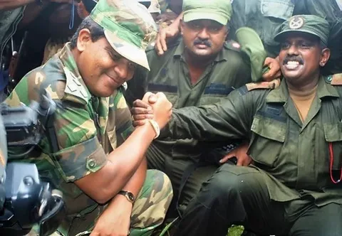 The image shows a group of military personnel in an outdoor setting. They are dressed in green camouflage uniforms and caps, typical of military attire. One of the individuals, identified in the hint as Shavendra Silva, is engaged in a friendly arm-wrestling gesture with another man. Both appear to be smiling, suggesting a moment of camaraderie or celebration. The background is filled with other military personnel, some capturing the moment with cameras, indicating that this could be an event or occasion of some significance. The lighting is natural, consistent with an outdoor environment. The mood of the image seems to be positive, with the individuals expressing a sense of unity and friendship. The focus is mainly on the central interaction between the two men, highlighting their relationship or camaraderie.