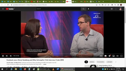 The image is a screenshot from a YouTube video titled "Facebook execs Sheryl Sandberg and Mike Schroepfer | Full interview | Code 2018," hosted on the Recode channel. It features two individuals, identified in the hint as Sheryl Sandberg and Mike Schroepfer, engaged in a conversation on stage. They are seated on red chairs against a backdrop with a mix of purple and blue lighting, suggesting a formal interview setting.
Sheryl Sandberg is dressed in a black top and is turned slightly towards Mike Schroepfer, showing an attentive posture. Mike Schroepfer is wearing a checkered shirt and glasses, facing Sheryl as they discuss a topic. The lighting highlights their profiles, creating a professional atmosphere. On the top right corner, there's a logo indicating the event is presented by the Alibaba Group.
Captions in the screenshot provide part of their conversation: "violence the numbers 86 what hate speech," and "it's 38 percent because it's harder." These subtitles suggest that they are discussing issues related to violence and hate speech, possibly in the context of social media moderation or policy.