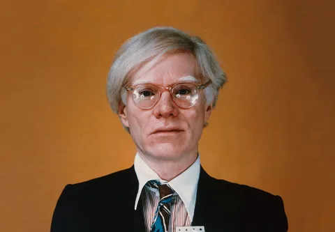 The image is a formal portrait of Andy Warhol. He is positioned against a plain, warm-toned background that contrasts with his attire. Warhol is dressed in a suit with a white shirt and a patterned tie, giving a professional appearance. His expression is neutral, and he looks directly at the camera, creating a sense of direct engagement with the viewer. Warhol is wearing clear glasses, and his light hair is neatly styled, adding to the composed and iconic look. The lighting is even, highlighting his features and providing a clear view of his face. The overall composition is straightforward, emphasizing the subject without any distracting elements in the background. This style is characteristic of traditional portrait photography, focusing entirely on the individual.