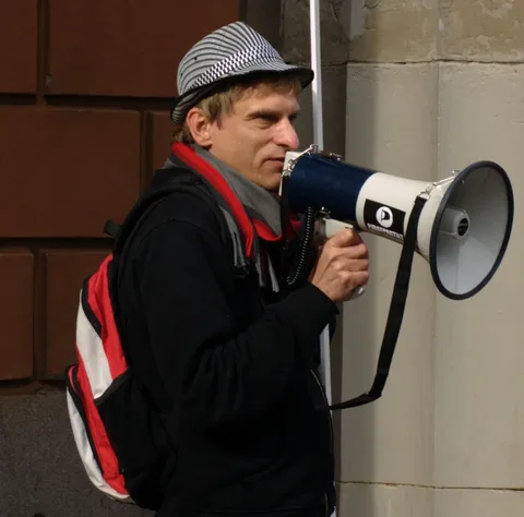 The image depicts an individual standing outdoors, possibly at a protest or public demonstration. The person is holding a blue and white megaphone, which features a sticker or logo. They are dressed casually in a black hoodie and a stylish, patterned hat. A red and white backpack is slung over their shoulder, and a red and gray scarf provides additional color and warmth. The background consists of a stone wall, indicating an urban setting. The individual appears to be actively speaking or shouting into the megaphone, suggesting a moment of engagement or advocacy. The lighting suggests a daytime scene, with natural light illuminating the subject. The mood conveyed is one of determination or activism, common in public speaking events or protests.