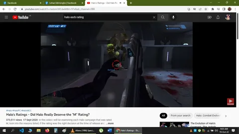 The screenshot is from a YouTube video titled "Halo's Ratings - Did Halo Really Deserve the 'M' Rating?" uploaded by the channel EBR. The video discusses the rating given to the Halo video game series, questioning whether it deserved its "M" (Mature) rating. In the image, a first-person view from the game Halo is shown, featuring an intense scene with a character engaging in combat against alien creatures in a sci-fi environment. The player’s hand is visible holding a weapon, aiming at an approaching enemy. There are clear signs of combat, with a red reticle indicating targeting and a "Reload" prompt on-screen as part of the gameplay interface. The environment is metallic and futuristic, with dim lighting and some blood visible on the floor. Key gameplay elements such as health and ammo counters are displayed in the upper left of the screen. The video had 373,511 views as of September 17, 2020. The screenshot also includes navigation elements from the YouTube interface, like the search bar and tabs for other open applications.