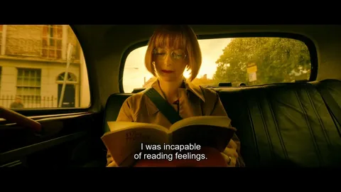 This image depicts a scene inside a car with a woman reading a book titled "The Prophet." The lighting is warm and casts an orange hue, suggesting it might be either early morning or late afternoon. The woman has short brown hair and is wearing glasses, which add an intellectual touch to her appearance. She is seated in the backseat of the car, and her seatbelt is visibly fastened across her chest. The background outside the car window is slightly blurred, indicating motion, with trees and a building passing by. The atmosphere inside the car feels introspective, enhanced by the subtitle "I was incapable of reading feelings," which suggests a theme of emotional disconnect or introspection. The woman appears focused and absorbed in her reading, contributing to a quiet, contemplative mood.