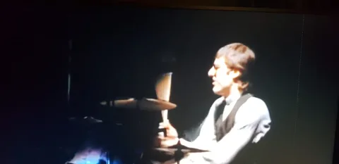 The image shows a drummer on stage, performing in what seems to be a concert setting. The lighting is dim, focusing primarily on the drummer, who is positioned to the right of the image. He is engaged in playing the drums, holding drumsticks with visible motion blur, suggesting dynamic action. He wears a light-colored shirt and a dark vest, giving him a formal appearance that contrasts with the energetic nature of his performance.
The background is dark, likely emphasizing the musician and the instruments in the foreground. The drum set is partially visible, with the cymbals catching some of the stage light. The overall mood is intense and focused, typical of a live musical performance.
Based on the hint, this scene appears to be from a performance by a drummer from Bruce Springsteen's band, possibly during a rendition of "Born in the USA". The setting suggests an intimate yet powerful concert atmosphere.