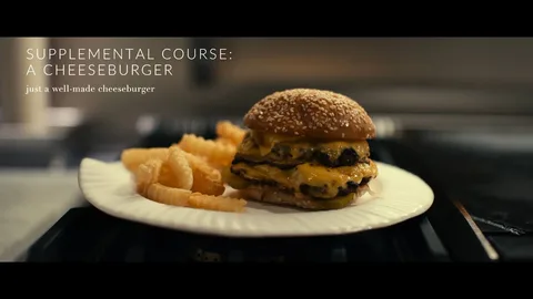 The image is a close-up from the film "The Menu," featuring a cheeseburger as the focal point. The cheeseburger is placed on a white plate alongside crinkle-cut fries. The burger consists of a sesame seed bun with layers of melted cheese and patties, giving a satisfying, appealing look. The simplicity and focus on the cheeseburger are highlighted by the text on the left side of the image, which reads "SUPPLEMENTAL COURSE: A CHEESEBURGER" followed by "just a well-made cheeseburger." The lighting is warm and inviting, emphasizing the juicy and freshly made appearance of the food. The setting seems to be indoors, likely in a kitchen or dining environment, consistent with the film's theme of culinary experiences. The composition draws attention to the meal, capturing the essence of a classic, well-prepared cheeseburger.