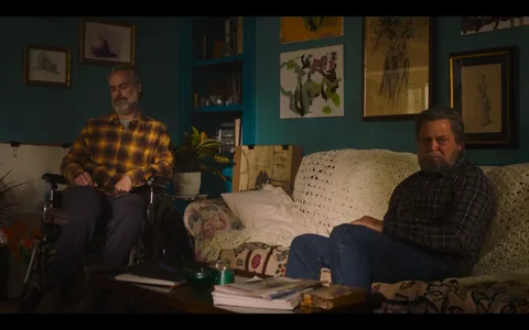 The scene is from "The Last of Us" featuring Nick Offerman. It takes place in a cozy, warmly lit living room adorned with various artworks on the teal walls. Two men are present: one is seated in a wheelchair wearing a yellow plaid shirt, and the other is sitting on a couch draped with a white crochet blanket, wearing a dark plaid shirt and jeans. Both men exhibit somber expressions, suggesting an emotional or serious conversation. The room is filled with personal touches like plants, framed pictures, and a table cluttered with papers and books, creating an intimate and lived-in atmosphere. The lighting is soft and natural, casting gentle shadows and highlighting the emotional weight of the scene.