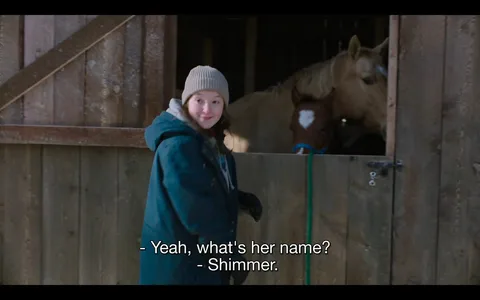 Ellie from "The Last of Us" is standing in a stable, looking back with a slight smile. She is dressed warmly in a green coat, gray hoodie, and beige knit hat, suggesting cold weather. Her expression is friendly and engaging, indicating a casual and pleasant interaction. In the background, two horses are visible inside the stable. One horse, notably a lighter color, stands next to a darker horse with a white mark on its face. The stable is constructed of wooden planks, contributing to a rustic atmosphere. The lighting is natural, suggesting daytime, and the scene feels serene and quiet. Subtitles at the bottom of the image read: "- Yeah, what's her name?" and "- Shimmer," indicating a conversation about one of the horses. This scene captures a moment of calm and connection with animals in the storyline.