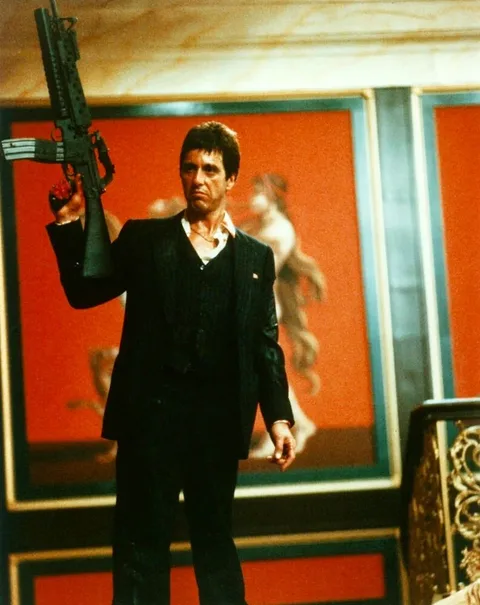 Tony Montana, portrayed by Al Pacino, stands prominently in this intense scene from the movie "Scarface." The character is holding a large firearm, exuding a sense of power and defiance. He is dressed in a dark pinstripe suit, which is slightly disheveled, adding to the intense atmosphere of the scene. His expression is fierce and determined, with a hint of defiance, capturing the iconic and intense nature of his character. The background features opulent decor with red hues and artwork, which contrasts with the tension of the moment. The lighting is warm, highlighting Montana's face and emphasizing the drama of the scene. This image captures a pivotal moment, showcasing the character's larger-than-life persona and the film's themes of crime and ambition.
