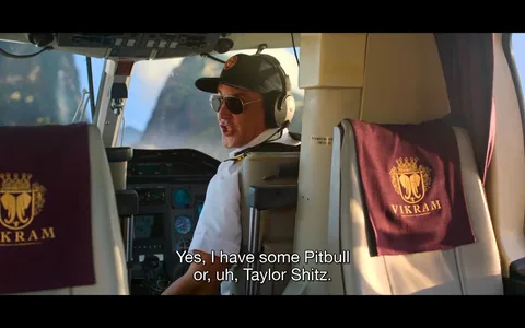 The image shows a scene inside a helicopter cockpit. A pilot, wearing a cap, sunglasses, and large headphones, is seated and facing slightly towards the back. He appears to be speaking with a playful or humorous expression. The pilot is dressed in a white shirt, possibly a uniform, which suggests he is on duty. In front of him, various controls and dials are visible, indicating the helicopter’s operational systems.
To the side, there is a seat covered with a maroon cloth bearing a crest and the name "VIKRAM," adding a touch of luxury or branding to the scene. The background through the cockpit windows displays a bright outdoor setting with blurred landscape views, suggesting movement or flight. The lighting in the cockpit is natural and bright, indicative of daytime. The scene has a comedic tone, as indicated by the subtitle, where the pilot humorously references musical artists.
- Captions / subtitles: "Yes, I have some Pitbull or, uh, Taylor Shitz."