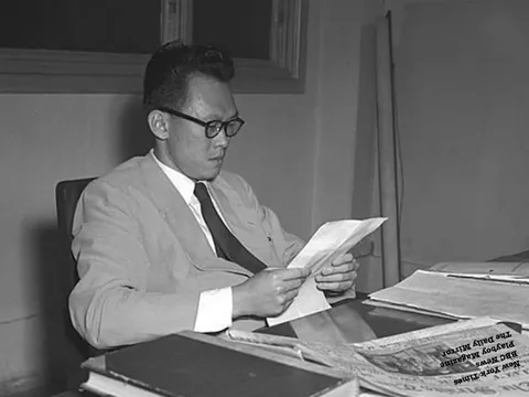 The black-and-white image depicts Lee Kuan Yew sitting at a desk in an office setting. He is absorbed in reading a document, which he holds with both hands. His demeanor is focused and serious, suggesting concentration on the contents. Lee Kuan Yew is dressed in a suit and tie, typical of professional attire, with glasses accentuating his intellectual appearance. The desk he is seated at is cluttered with various papers and books. To the right, several newspapers and publications are visible, including titles like "New York Times," "BBC News," "Playboy Magazine," and "The Daily Mirror," indicating a diverse range of reading material. The background is simple, with a window partially visible, adding depth to the office environment. The monochromatic palette of the image gives it a historical feel, capturing a moment that seems significant in the context of Lee Kuan Yew's life and work.