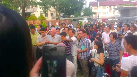 The image captures an outdoor scene at a tree planting ceremony in Holland Village, Singapore. A crowd gathers around, with Lee Kuan Yew prominently featured in the center. He is surrounded by people who appear attentive and engaged, indicating a significant moment. The scene is bustling, with attendees of different ages and backgrounds, some holding cameras and phones, suggesting they are either documenting the event or just participating as onlookers.
Lee Kuan Yew is wearing a light-colored, checkered shirt, and appears poised and focused on the event. The atmosphere seems warm and communal, with people standing closely together, indicative of a shared public moment. In the background, residential buildings and greenery are visible, providing a typical urban Singaporean backdrop. The lighting is natural, suggesting it's either morning or late afternoon.
The image captures a moment of both historical and cultural significance, reflecting Lee Kuan Yew's connection with the community and his legacy in Singapore.