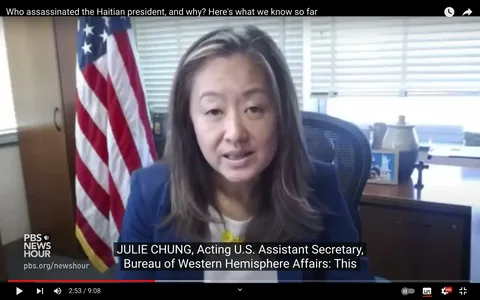 The image is a video frame from a YouTube video titled "Who assassinated the Haitian president, and why? Here's what we know so far" on the PBS NewsHour channel. In the frame, Julie Chung, identified as the Acting U.S. Assistant Secretary for the Bureau of Western Hemisphere Affairs, is speaking during an interview. She is seated in an office setting with an American flag prominently displayed behind her, suggesting a formal government environment.
Julie Chung is wearing a professional outfit, with a navy blazer over a white top. Her expression is earnest, reflecting the serious nature of the subject matter. There is a wooden desk in the foreground with various items, including a framed photograph and a decorative vase. The lighting is neutral, giving the scene a clear and professional appearance.
The on-screen text includes the PBS NewsHour logo and website, along with subtitles that capture Julie Chung's role and her speech. The video discusses the assassination of the Haitian president, a topic of significant geopolitical interest.