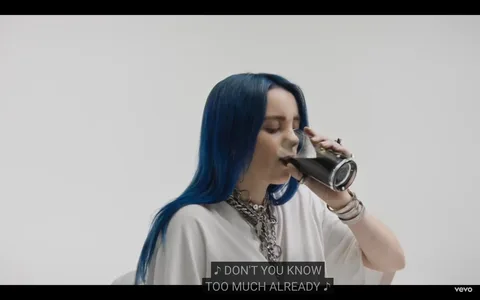 The image is a frame from a music video featuring Billie Eilish. She is seated indoors, drinking from a glass filled with a dark liquid, possibly soda or coffee. Billie has long, blue hair and is wearing a casual white outfit accented with a chunky silver chain necklace and multiple bracelets. Her eyes are closed as she takes a sip, suggesting calmness or introspection. The background is simple and white, focusing attention on her. Captions at the bottom of the frame read, "DON'T YOU KNOW TOO MUCH ALREADY," adding a lyrical element from the video. The lighting is clear and bright, enhancing the clean and minimalistic aesthetic typical of Billie Eilish's music videos.