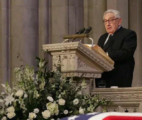 Henry Kissinger is depicted speaking at a podium inside a formal setting, likely a cathedral or large church given the architectural style and the presence of tall stone pillars in the background. The podium is ornate, crafted from stone or a similar material, with detailed carvings. Positioned prominently in front of him are two microphones, suggesting he is delivering an important speech or eulogy.
Kissinger is wearing a dark suit with a white shirt and a dark tie, adding to the somber and formal atmosphere of the scene. His expression is serious and focused, consistent with the gravity of the occasion. In the foreground, a large arrangement of white flowers, possibly lilies and roses, adds a touch of solemnity and elegance, indicating a ceremonial or memorial event. In front of the podium, the edge of a flag-draped casket is visible, reinforcing the likelihood of a funeral setting. The lighting is soft yet sufficient to highlight Kissinger and the podium, creating a respectful and reverent ambiance.