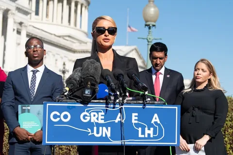 Paris Hilton is at the center of this outdoor press conference, standing in front of a building that resembles the U.S. Capitol. She is wearing a black outfit and large sunglasses, speaking into multiple microphones from various media outlets. The microphones are on a podium with a blue sign displaying partially obscured text, "CO GRE A E A UN LH," which appears handwritten in white.
To her right, a man in a suit and polka dot tie holds a green folder titled "Away From Home: Youth Experiences of Youth-Based Placement in Foster Care." On her left, another man in a suit with a red tie stands with a serious expression. Beside him, a woman in a black dress, appearing to be pregnant, stands attentively.
The scene suggests an advocacy event, likely related to youth and foster care, given the visible document. The weather is clear and sunny, indicated by the bright lighting and blue sky in the background. The American flag is visible, adding to the formal and political atmosphere of the event.