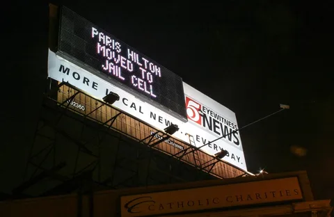 The image shows a large outdoor billboard at night, with a prominent electronic display at the top. The billboard is located outdoors, illuminated against the dark night sky. The electronic display features scrolling text that reads, "PARIS HILTON MOVED TO JAIL CELL," suggesting a news update regarding Paris Hilton. Below this, the billboard advertises "5 Eyewitness News" with the "abc" logo visible, indicating that this is a news broadcast station. The words "MORE LOCAL NEWS. EVERY DAY." are prominently displayed beneath the electronic text. The billboard is situated above a building marked "CATHOLIC CHARITIES," suggesting a location in a populated urban area. The lighting highlights the billboard's structure and text clearly, emphasizing the news headline. The scene captures the contrast between the bright lights of the billboard and the dark surroundings, drawing attention to the news being broadcast.