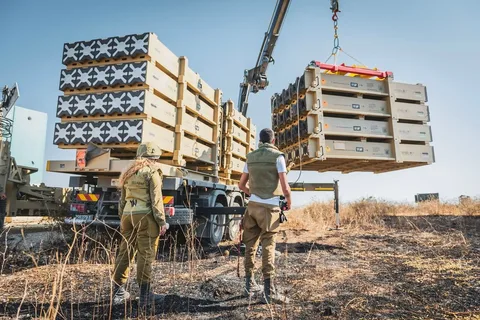 In the image, two individuals in military attire are overseeing the loading of large crates onto a vehicle. The crates are being lifted by a crane, indicating a logistical or operational military task. The setting appears to be outdoors, with dry grass underfoot and a clear blue sky above, suggesting a warm and arid environment.
The military personnel are wearing uniforms with protective gear, including helmets and vests, indicating safety precautions during the operation. One person is facing the vehicle, while the other looks at the crane, both observing the process closely. The crates being moved have visible labels, including the word "ANALOG" and the number "17," suggesting they contain standardized equipment or materials.
The vehicle and crane are positioned on a slightly uneven terrain, with the equipment suggesting a temporary or field operation. The scene is well-lit by natural sunlight, adding to the clarity of details such as the color of the uniforms and the markings on the crates. There is a sense of coordination and control as the personnel manage the equipment, indicating a routine procedure in a military context.