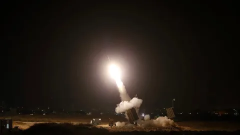 The image shows a night-time scene of a missile being launched from a military defense system setup. The environment is dark, with the launch creating a bright, fiery glow against the night sky. The missile is ascending at a steep angle, leaving a trail of smoke and light behind. The launcher is positioned on the ground, surrounded by dry grass, and emits a cloud of smoke as the missile is fired. In the background, the silhouette of a city skyline with scattered lights can be seen, suggesting the location might be near an urban area. The scene conveys a sense of urgency and precision, characteristic of military operations. The image captures the dramatic contrast between the bright launch and the dark surroundings, emphasizing the power and technology involved in the defense system.