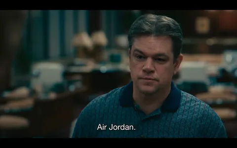 The image is a still from the film "Air," featuring a character played by Matt Damon. The setting is an indoor office environment, indicated by the presence of blurred office furniture and decor in the background. The lighting is soft and ambient, contributing to a serious and contemplative mood. Matt Damon is wearing a patterned polo shirt with a collar, in shades of blue, and his expression appears thoughtful and focused. The subtitle at the bottom of the image reads "Air Jordan," suggesting a conversation topic related to the famous basketball brand. The scene seems to center on a discussion or decision-making process, typical of a business or corporate context. The camera angle is at eye level, which helps to engage viewers with the character's perspective. Overall, the composition indicates a pivotal moment in the narrative of the film "Air."