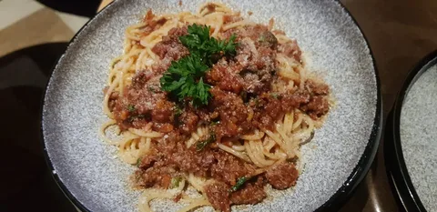 The image features a plate of spaghetti topped with a rich, meaty sauce, likely a classic spaghetti Bolognese. The pasta is neatly arranged on a textured, gray plate, creating a pleasing contrast with the reddish-brown sauce. The sauce appears to be made with ground meat and is garnished with a few sprigs of fresh parsley, adding a touch of green to the dish. The pasta is sprinkled with grated cheese, perhaps Parmesan, which complements the savory flavors of the dish. The lighting is warm, highlighting the textures and making the food look appetizing. The background is slightly out of focus, suggesting an indoor dining setting, possibly a restaurant or a home kitchen. The overall presentation is inviting and suggests a hearty, homemade meal.