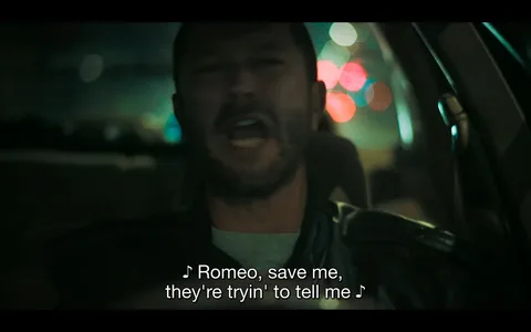 The image depicts a scene from a video or film where a man is sitting in a car at night, seemingly singing along to a song. The lighting is dim, with colorful bokeh lights visible through the car's window, suggesting a city setting. The man appears to be wearing a leather jacket and a neutral expression, though his mouth is wide open, indicating he might be passionately singing or speaking. The frame has a moody and atmospheric feel, with the focus primarily on the man's face. Captions at the bottom of the image read, "♪ Romeo, save me, they're tryin' to tell me ♪," which hints at the lyrics of the song being played or sung. The setting inside the car is intimate and close-up, emphasizing the emotional connection to the music.