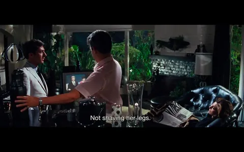 The image is a scene from the film "The Graduate." It features two men and a woman in an indoor living room setting. One man, seen in profile, is wearing a light-colored suit and is likely engaged in conversation with another man facing away from the camera, wearing a pink shirt. They are near what appears to be a bar area with various glassware and an ice bucket. The background is filled with lush greenery visible through large windows, suggesting an upscale and possibly modern home environment.
To the right, a woman lounges on a black leather chair, holding a magazine, adding a relaxed, casual vibe to the setting. The lighting is soft, with a mix of natural and ambient illumination, highlighting a cozy yet elegant atmosphere. The subtitle "Not shaving her legs." suggests a casual, perhaps humorous conversation is taking place. The overall mood seems relaxed, with an air of sophistication typical of the film's aesthetic.
- Captions / subtitles: "Not shaving her legs."