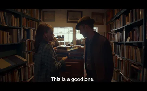 ```
The image depicts a scene in a cozy bookstore. Shelves filled with books line the left and right sides of the image, creating a warm, inviting atmosphere. In the foreground, a woman and a man are engaged in conversation. The woman, dressed in a plaid coat, is holding a book and appears to be discussing it with the man, who is wearing a hat and casual jacket. Their posture suggests a friendly interaction, possibly sharing recommendations or discussing literature. In the background, a window lets in soft daylight, illuminating the room. The lighting and colors contribute to a nostalgic, intimate mood typical of small bookstores. Above them, framed pictures hang on the wall, adding to the vintage charm of the setting. The subtitle at the bottom reads, "This is a good one," indicating a positive opinion about a book.