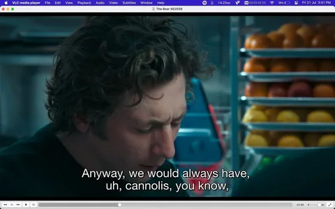 The image is a screenshot from the show "The Bear," specifically from Season 2, Episode 8, as indicated by the title text at the top of the screen. The visual is taken from a scene set indoors, likely in a kitchen environment. In the foreground, a person with curly hair is looking downward, possibly speaking or reflecting, with a serious or contemplative expression. The background features several racks filled with fruit, adding a vibrant touch with oranges and yellows. The lighting is soft and natural, typical of a kitchen setting, emphasizing the character's thoughtful demeanor. The interface elements indicate that the media is being played on VLC media player. The subtitles at the bottom read, "Anyway, we would always have, uh, cannolis, you know," suggesting a personal or nostalgic conversation. The overall mood of the scene appears introspective or reminiscent.