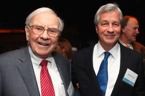 Warren Buffet and Jamie Dimon are pictured together in an indoor setting, likely at a formal event or gathering. Warren Buffet is on the left, wearing a dark suit with a white shirt and a red striped tie. He has a friendly expression and is looking directly at the camera. Jamie Dimon is on the right, dressed in a dark suit with a white shirt and a blue tie. He is smiling, and a name tag on his chest reads "Jamie Dimon" with "FORTUNE" visible below it. The background is somewhat dimly lit, with other people present, suggesting a social or professional event. The attire and expressions of the individuals convey a formal yet congenial atmosphere.