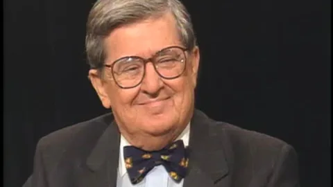 The image features AM Rosenthal in a studio setting. He is wearing a dark suit with a light-blue shirt and a patterned bow tie, giving off a formal and polished appearance. His expression is warm and approachable, with a slight smile, suggesting a relaxed and conversational mood typical of an interview or discussion. The background is a plain black, which helps to focus attention on Rosenthal himself. His hair is gray and neatly styled, and he is wearing large, round eyeglasses. The lighting is soft and even, highlighting his facial features without creating harsh shadows. The overall composition is centered, with Rosenthal's head and shoulders prominently displayed, indicating this is likely a still from a television interview or a similar on-camera appearance.
