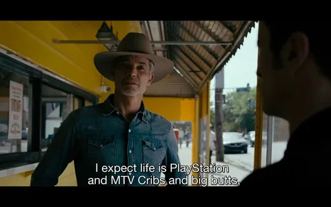 The image depicts an outdoor scene with two individuals engaged in conversation. One person, wearing a wide-brimmed hat and a denim shirt, stands in front of a brightly painted yellow building. His expression appears thoughtful as he addresses the other person. The building has a corrugated metal overhang, casting a shadow over the scene, suggesting a sunny day. A poster with text and pricing is visible on the left, indicating a commercial setting, possibly a diner or small restaurant. The background shows a street with a few vehicles and trees, providing an urban or suburban context. The subtitle at the bottom reads, "I expect life is PlayStation and MTV Cribs and big butts," adding a touch of humor and modern cultural reference to the dialogue. The overall mood is casual, with a focus on everyday life and interactions.
- Captions / subtitles: "I expect life is PlayStation and MTV Cribs and big butts,"