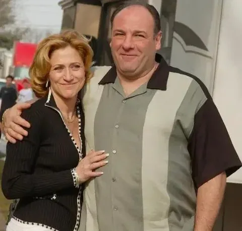 In this image, James Gandolfini is seen posing outdoors with a woman. The setting appears informal, likely outside a trailer or building, suggesting it might be on a film or TV set. James Gandolfini is wearing a short-sleeve, button-up shirt with a color block design in shades of green, gray, and black. He has a relaxed expression and is smiling gently while his arm is around the woman, indicating a friendly or collegial relationship.
The woman, who is also smiling, is wearing a black blouse with white trim and has her arm around Gandolfini. Her hair is styled in soft waves, and she appears content and comfortable in the setting. In the background, there is a large vehicle, possibly a trailer, and blurred figures or objects that hint at a bustling environment typical of a production set. The lighting is natural, suggesting it is daytime, and the mood is casual and pleasant.