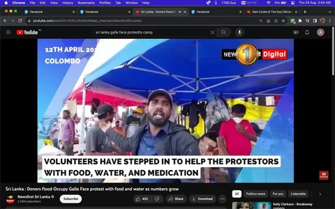 The image is a screenshot from a YouTube video titled "Sri Lanka : Donors flood Occupy Galle Face protest with food and water as numbers grow," published by Newsfirst Sri Lanka. The scene captures a protest camp, likely at Galle Face in Colombo, Sri Lanka, on April 12, 2022, as indicated by the on-screen text. The foreground features a man speaking energetically, possibly addressing the camera or other individuals in the vicinity. He wears a cap and a jacket, suggesting casual attire suitable for outdoor activities.
Surrounding him are several tents in blue and red, under which other individuals move about, some engaging with supplies. One person in a red shirt and mask appears to be organizing or distributing items. The setting is vibrant, with bright colors from the tents and various supplies visible in the background, indicating active volunteer involvement. The subtitle highlights that volunteers are providing food, water, and medication to support the protestors. The atmosphere is bustling with activity, reflecting the community's collective effort in aiding the demonstrators.