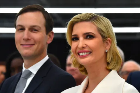 Jared and Ivanka are prominently featured in this image, which appears to be taken at an indoor event. They are both smiling warmly, suggesting a positive and relaxed atmosphere. Ivanka is wearing a white outfit, with her hair styled in a sleek, short bob, complemented by elegant earrings. Her makeup is polished, emphasizing her eyes and smile. Jared, standing beside her, is dressed in a dark suit with a white shirt and tie, exuding a formal yet approachable demeanor. The lighting is bright and even, highlighting their faces against a blurred background of other attendees. The composition centers them, indicating they might be significant figures at this gathering. The overall mood is one of composure and professionalism.
