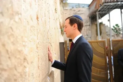 Jared is depicted in a solemn moment of reflection at the Western Wall, a significant religious site in Jerusalem. He is wearing a dark suit and a kippah, which is customary for men at this location, indicating respect and adherence to tradition. Jared is standing in profile, gently placing his hand on the ancient stones of the Wall, a common practice symbolizing a connection to faith and history. The background shows the characteristic large stones of the Wall, which are weathered and imbued with historical significance. The lighting is natural and soft, suggesting that this is taking place during the day. The scene captures a quiet, introspective moment amid the bustling atmosphere typically found at this site, emphasizing personal spirituality and contemplation.