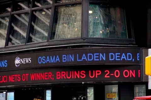 The image depicts a news ticker on the exterior of a building displaying breaking news headlines. The prominent text reads "OSAMA BIN LADEN DEAD," indicating a major news announcement about the death of Osama bin Laden. This text is in blue, making it stand out against the black background of the ticker. Below, in red text, a sports update reads, "BRUINS UP 2-0 ON P," which likely pertains to a playoff series involving the Boston Bruins.
The building is identified as part of ABC News, with their logo visible on the ticker. The setting appears to be outdoors, possibly in a bustling urban area, given the architectural style and the presence of large windows reflecting the surrounding environment. The structure suggests a modern media center, likely in a well-known city location, such as Times Square. The lighting is natural, and the image captures part of a street sign or traffic light pole in the bottom right corner, adding to the urban context. The overall mood is serious and urgent, typical of breaking news scenarios. The design and placement of the ticker are intended to attract public attention quickly and convey important information efficiently.